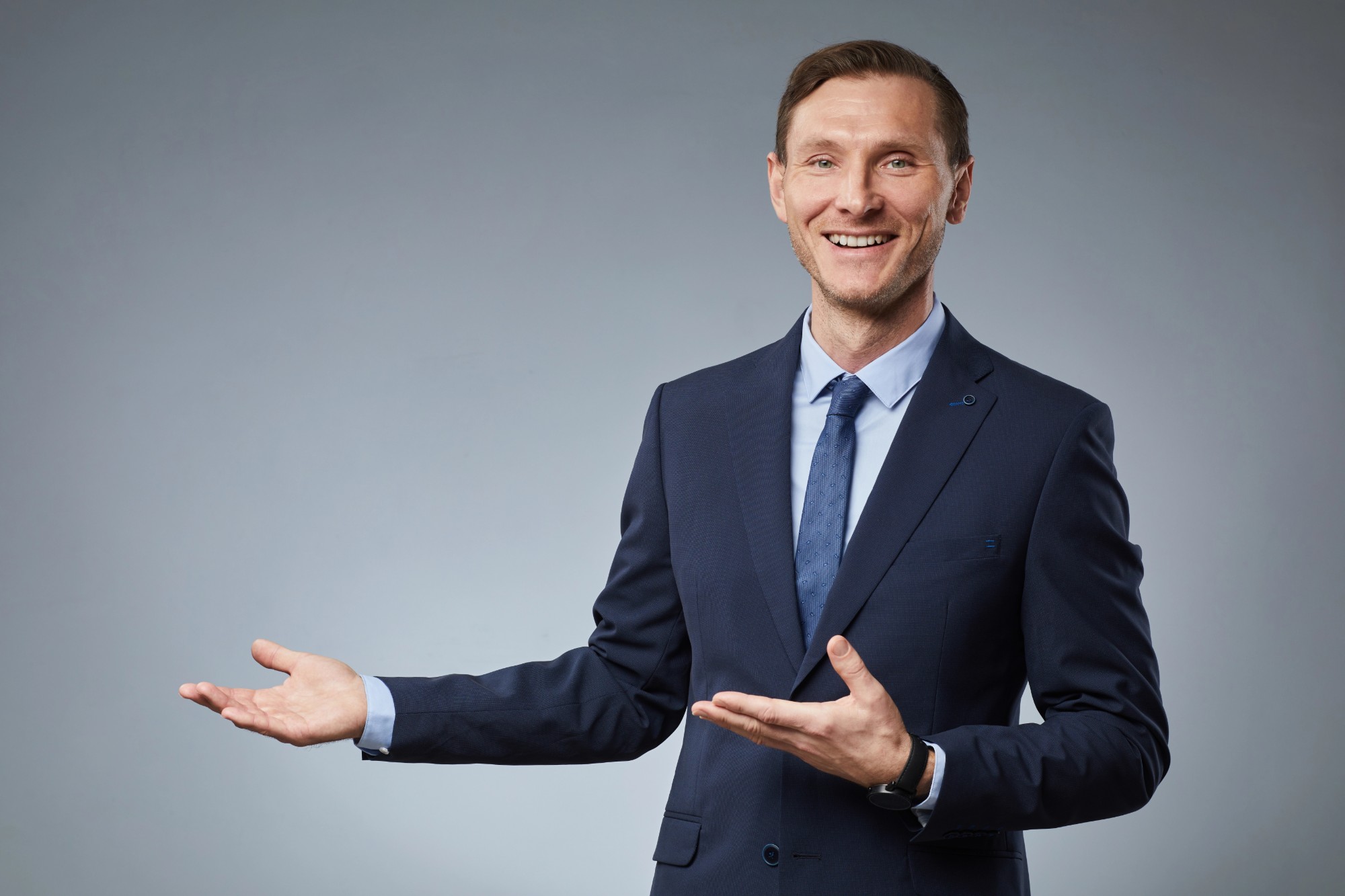 Presenter in a suit gesturing to camera against a grey studio backdrop